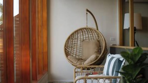 Une chaise en osier avec un coussin, une petite table ronde et un oreiller à rayures, dans une pièce au plancher en bois et dotée d’un grand miroir.