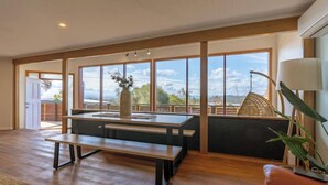 Une pièce spacieuse avec un banc en bois et une table, de grandes fenêtres donnant sur l’extérieur et une plante en pot.