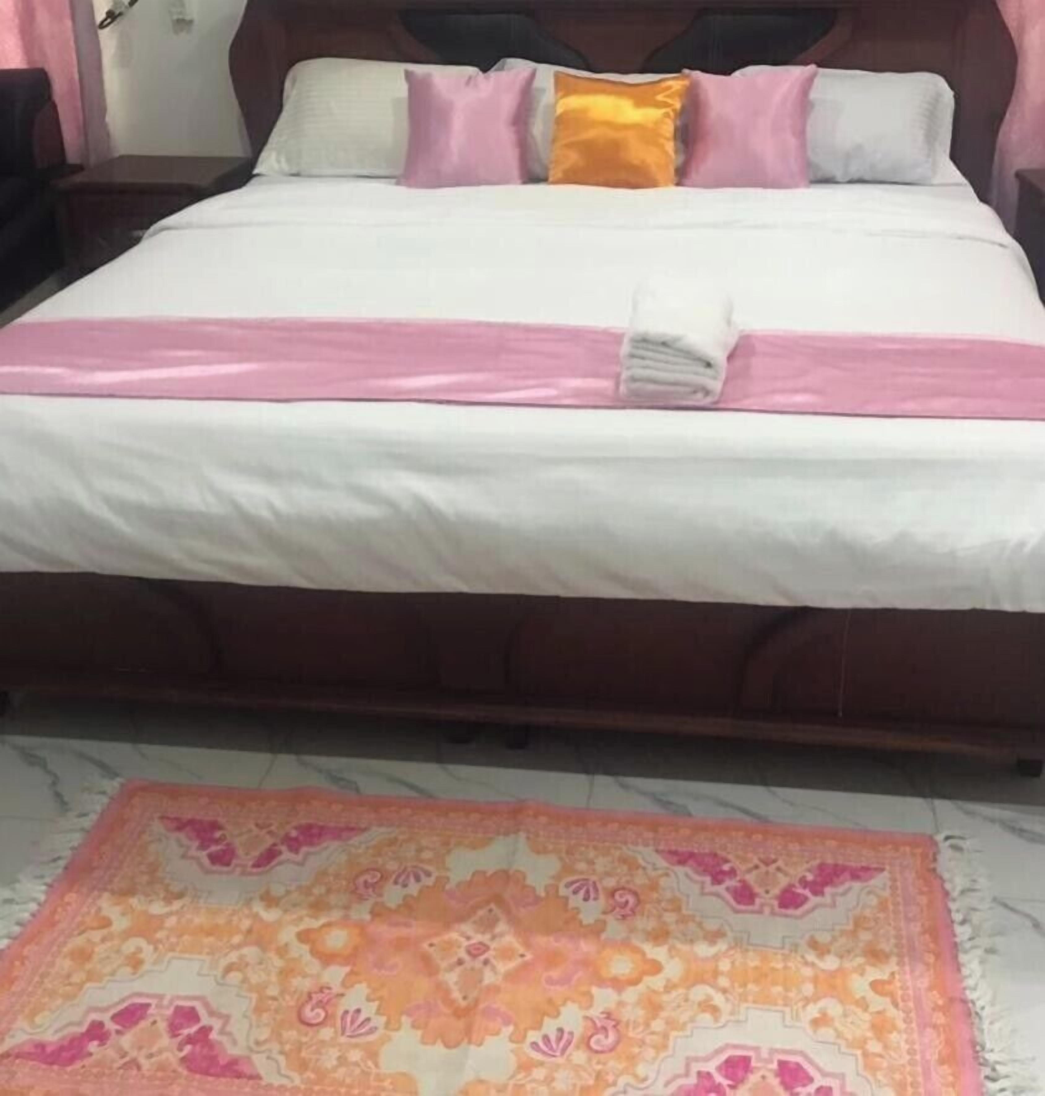 A bed with a white and pink bedspread, a pink blanket, and a folded white towel. Below the bed, there is a patterned rug with orange and pink colors.