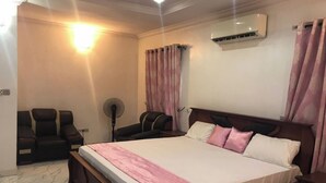 Une chambre avec un grand lit, deux chaises, un ventilateur sur pied et un appareil de climatisation.