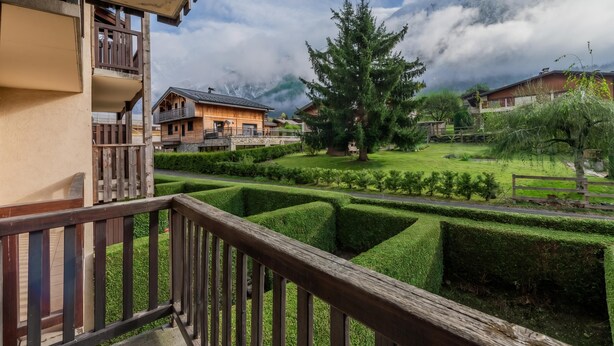 Ein Chalet mit Holzbalkon, ein gepflegter Garten mit Hecken und eine Bergkulisse im Hintergrund.