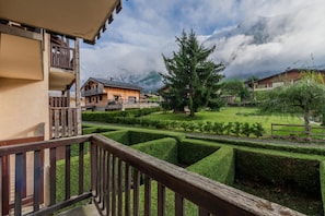 A chalet-style building with a wooden balcony, a well-manicured garden with hedges, and a mountain backdrop.
