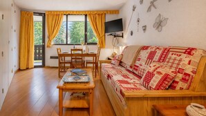 Ein gemütliches Zimmer mit einer Holzdavonia, einem Esstisch, Stühlen, einem an der Wand montierten Fernseher und einem Fenster mit Vorhängen.