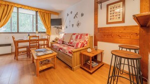 Ein gemütliches Zimmer mit einer Holzcouch, einem Esstisch mit Stühlen, einem Fernseher und einer Holztresorecke.
