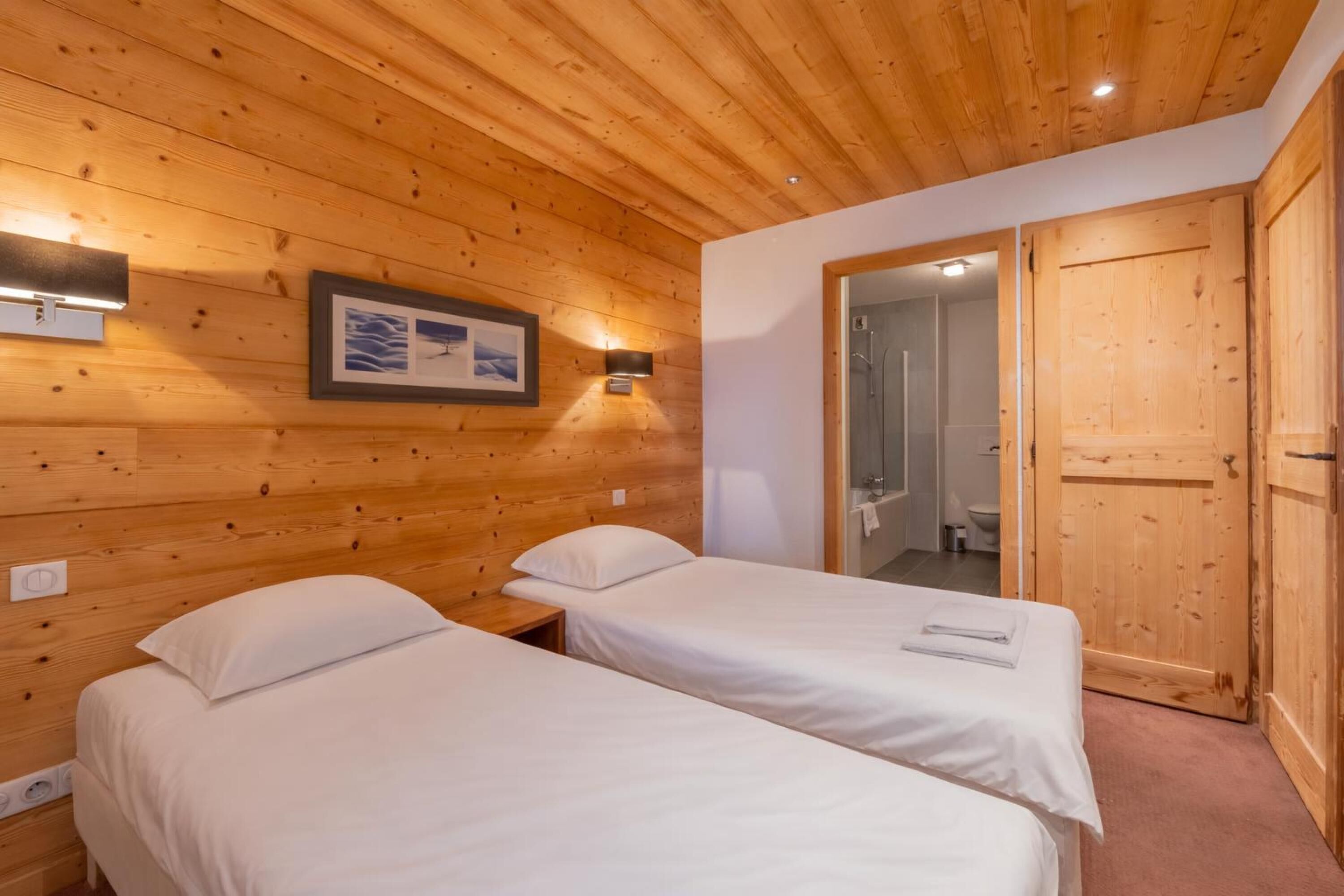 A wooden-paneled room with two single beds, a framed picture on the wall, and a door leading to a bathroom.