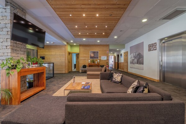 A modern lobby with a wooden ceiling, a flat-screen TV mounted on a stone wall, a sofa, a coffee table, and a reception desk.