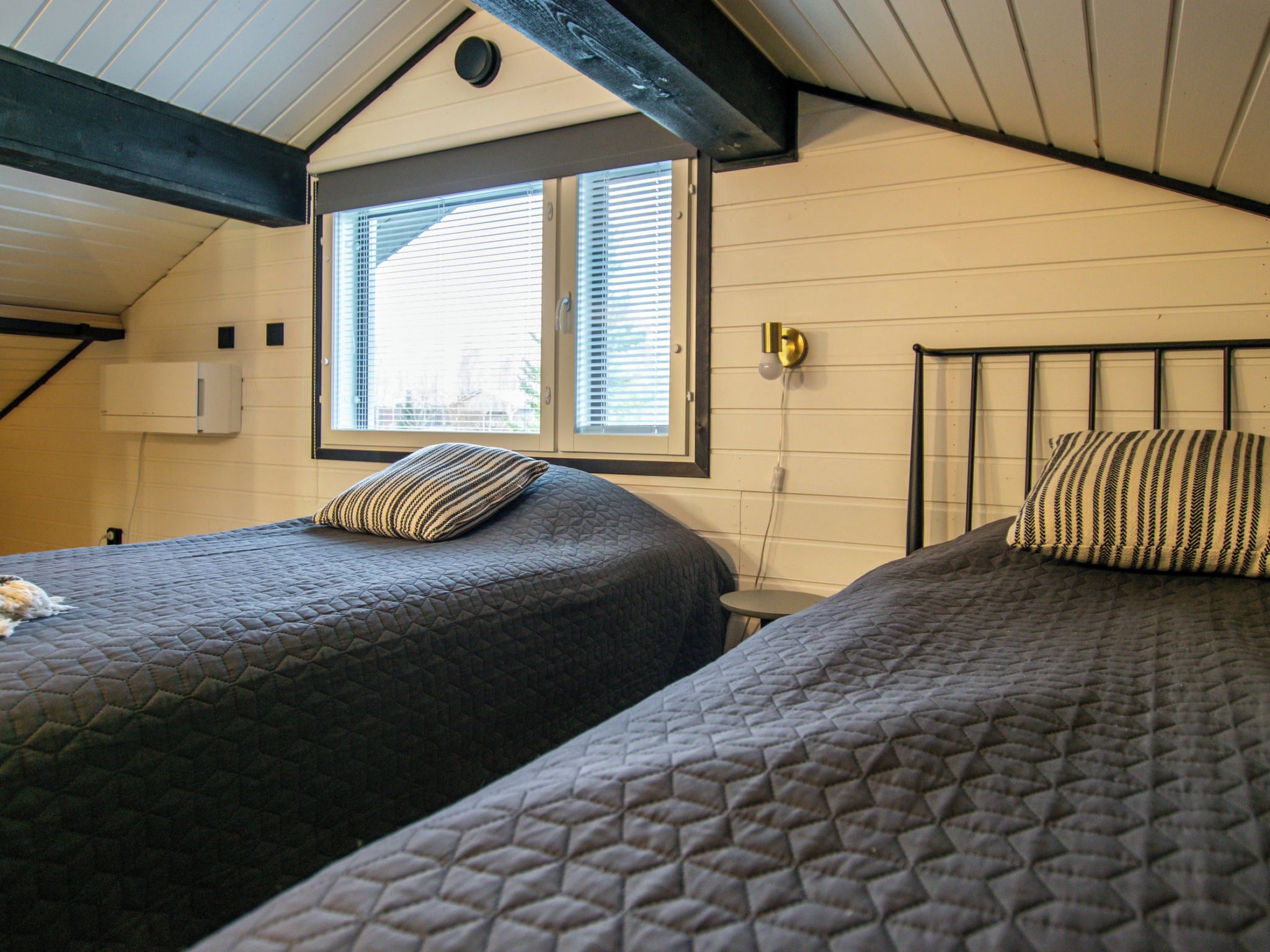 A room with two beds, a window with blinds, a wall-mounted light fixture, and a ceiling with exposed beams.