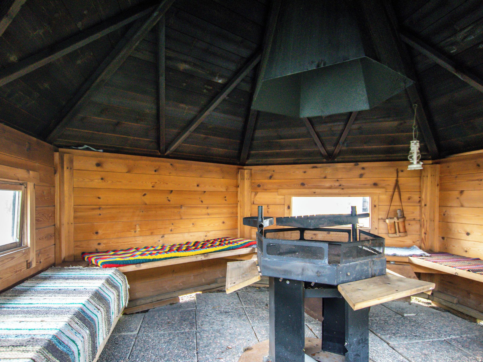 L’intérieur d’une cabane avec un plancher en bois, un banc, une table recouverte d’un tapis tressé et une cheminée.