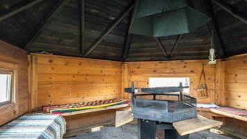 L’intérieur d’une cabane avec un plancher en bois, un banc, une table recouverte d’un tapis tressé et une cheminée.