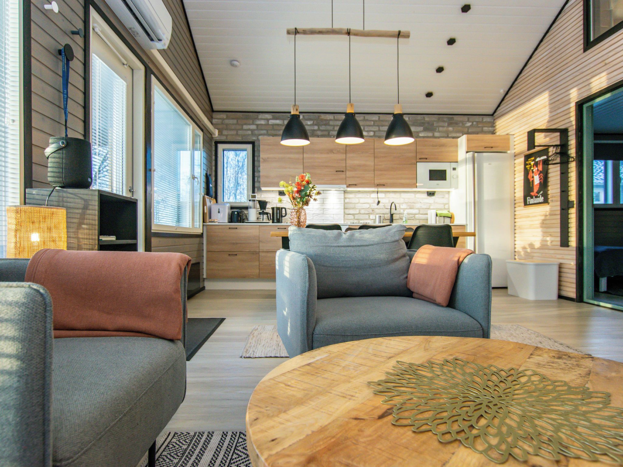 A modern kitchen with wooden cabinets, a dining area with a round wooden table, and a grey sofa with a throw pillow.