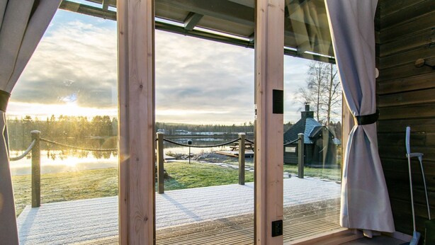 Ein Zimmer mit großen Schiebeg lassfenstern und Zugang zu einer Holzterrasse mit Blick auf einen See.