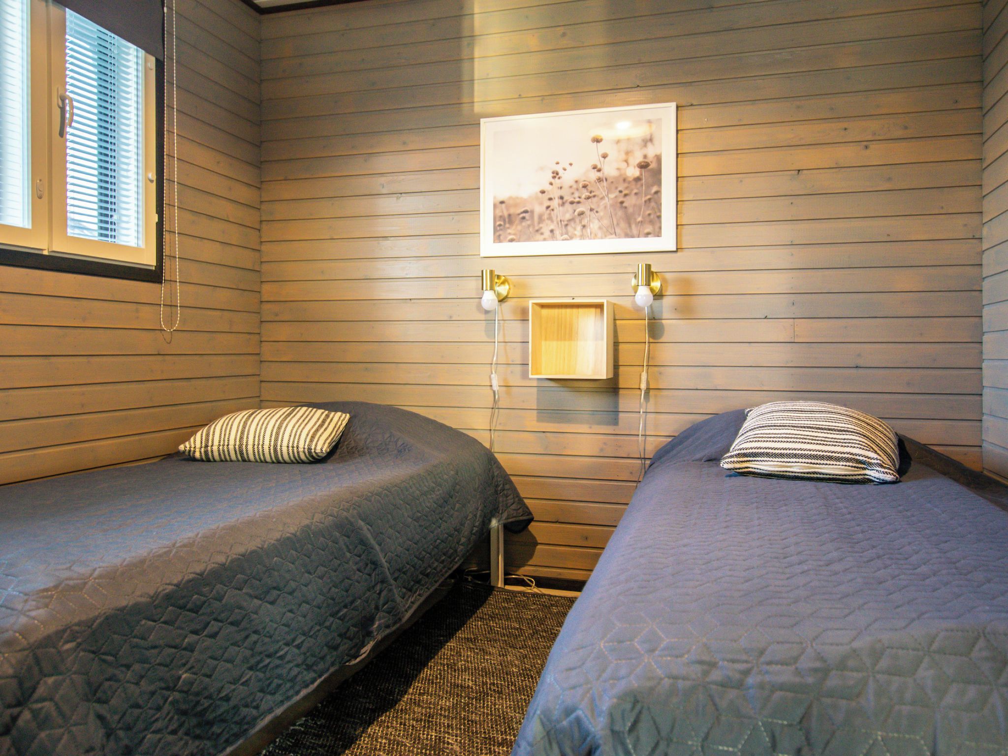 A room with two single beds, wooden walls, a framed picture, and a small table with a lamp.