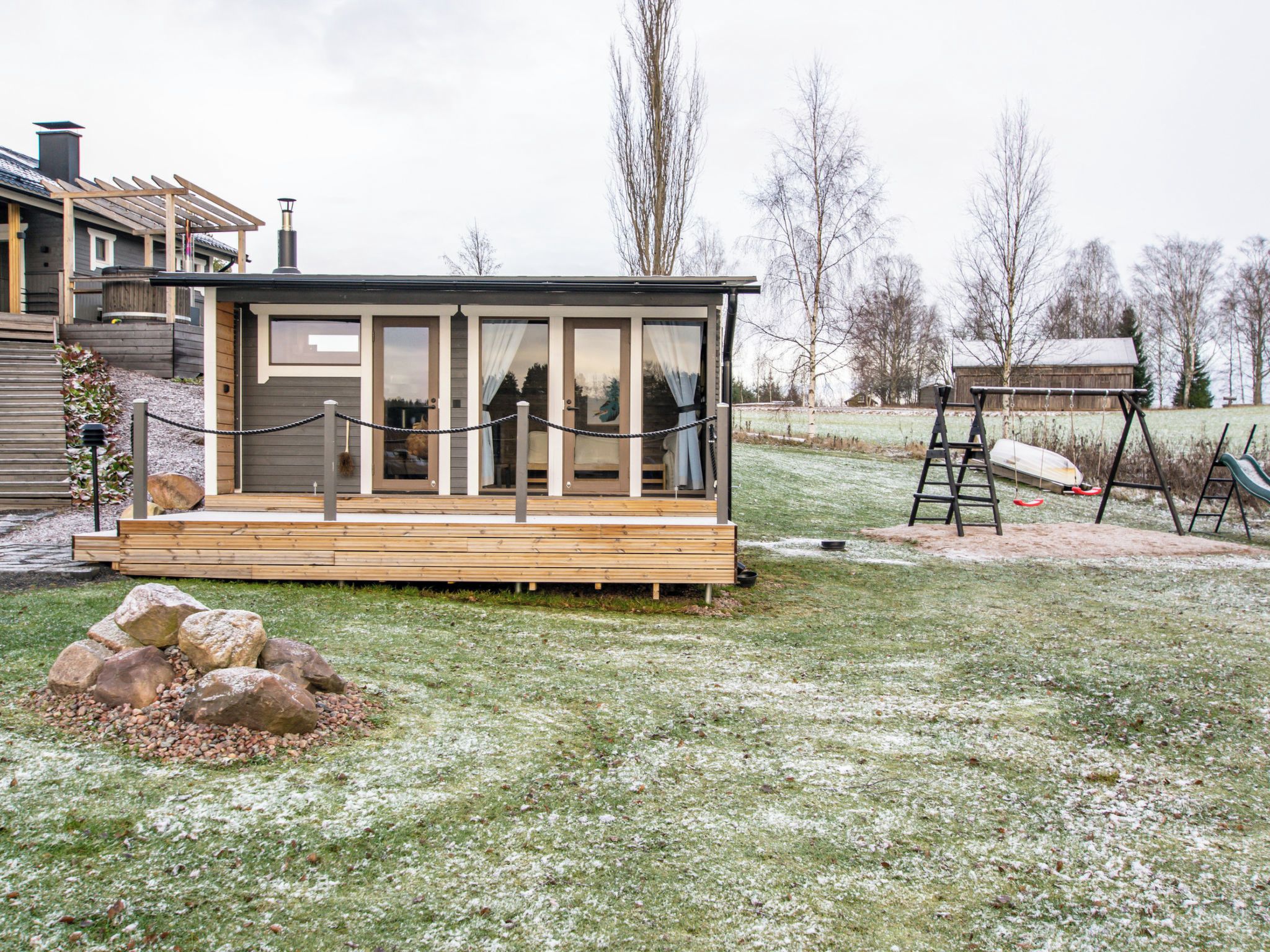 A small cabin with a wooden deck, a glass door, and a fenced area with a swing set.