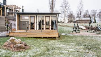 A small cabin with a wooden deck, a glass door, and a fenced area with a swing set.