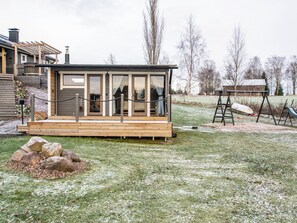 Une petite cabane avec une terrasse en bois, une porte vitrée et un espace clos comprenant une balançoire.