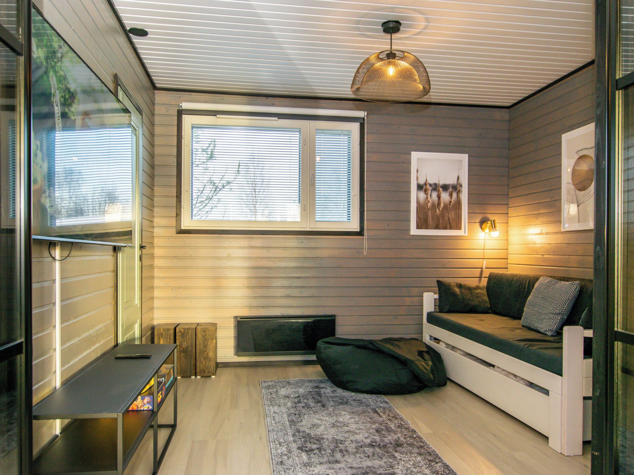 A cozy room with a bed, a small table, a window with blinds, and a wall-mounted light fixture.