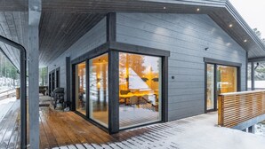 Una moderna baita con una terrazza in legno e ampie porte vetrate che danno su un interno con camino e zona salotto.