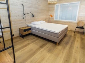 Een slaapkamer met een bed, nachtkastje, lamp, raam en houten vloer.