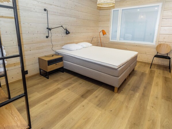 A bedroom with a bed, bedside table, lamp, window, and wooden floor.