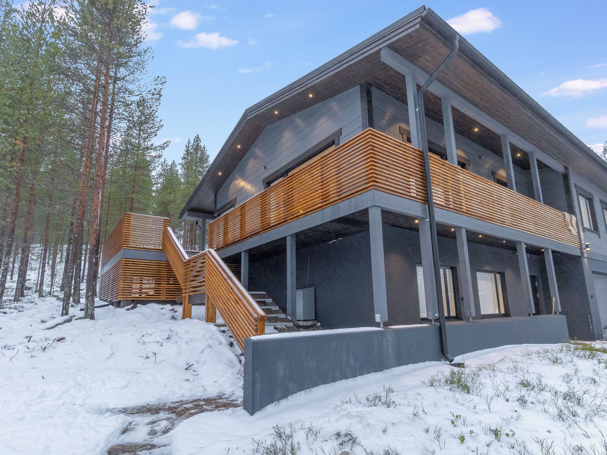 Un bâtiment moderne à plusieurs étages, doté d’un escalier en bois menant à un balcon, entouré de neige et d’arbres.