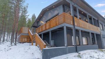 A modern multi-story building with a wooden staircase leading to a balcony, surrounded by snow and trees.