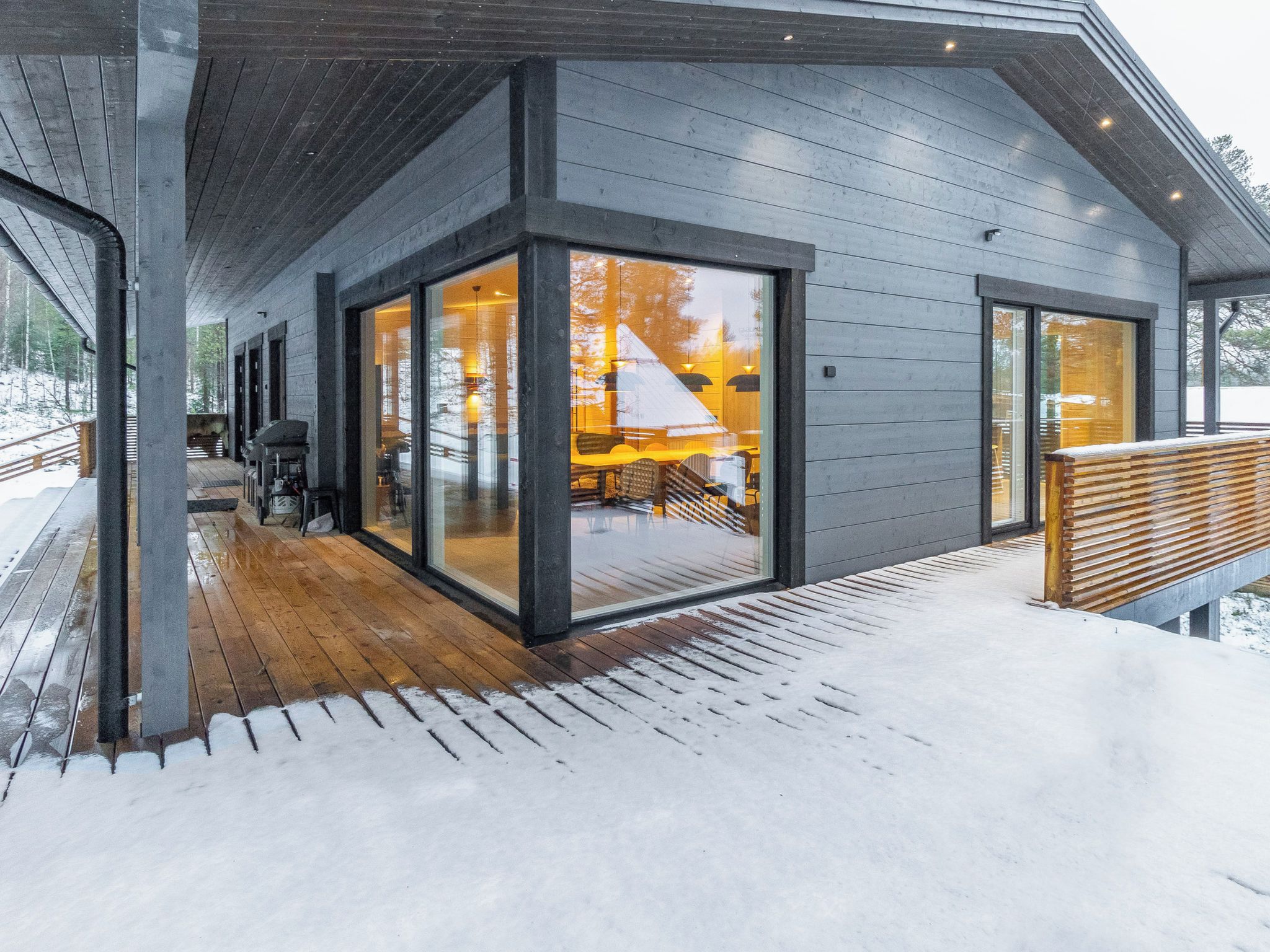 Un edificio moderno con amplios ventanales, una terraza de madera y suelo cubierto de nieve.