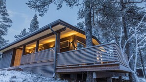 Una cabaña moderna con terraza de madera, ventanas amplias y cálida luz interior.