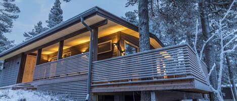 A modern wooden cabin with a balcony, surrounded by snow-covered trees.