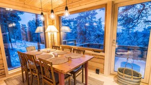 Un interior de madera con una mesa de comedor y sillas, ventanas amplias con vistas nevadas y lámparas colgantes.