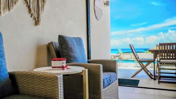 Ein Zimmer mit Korbmöbeln, einem kleinen Tisch mit einer roten Geschenkbox und Blick auf den Strand durch eine Glastür.
