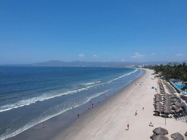 Ein Strand mit blauem Himmel, sandigem Ufer und einer Reihe von Reetdachschirmen.