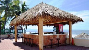 Un espace bar extérieur doté d’un toit de chaume, offrant une vue sur l’océan, avec des tabourets en bois et un sol pavé de briques.