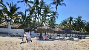 Une plage avec des parasols en chaume, des palmiers et un panneau indicateur.