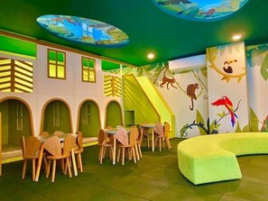 Un espace de jeux pour enfants avec un toboggan, des chaises en bois, une table et un banc vert rembourré.