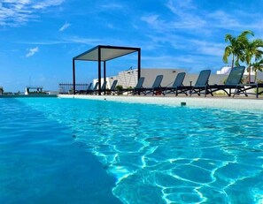 Piscina azul cristalina con sillas de descanso y una zona sombreada, rodeada de edificios y palmeras.
