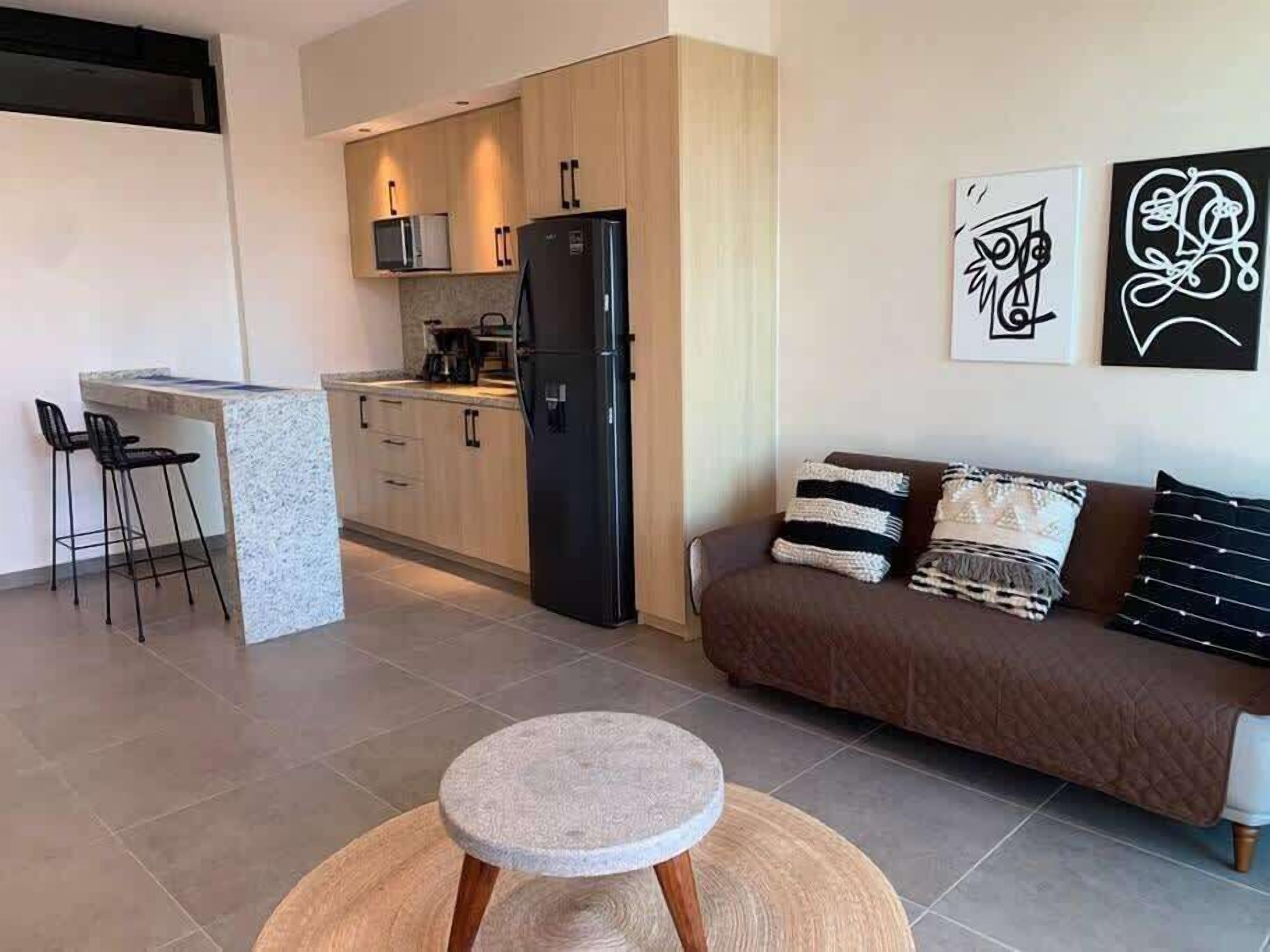 A modern kitchen with a refrigerator, microwave, and bar stools. There is a round stone table in the foreground and a brown sofa with patterned cushions.