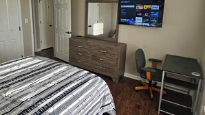 Un dormitorio con cama, cómoda, escritorio, silla y televisor montado en la pared que muestra la interfaz de una plataforma de streaming.