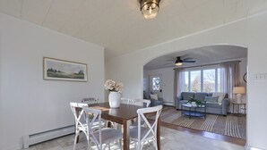 A dining area with a wooden table and white chairs, a white vase with flowers, and a painting on the wall.