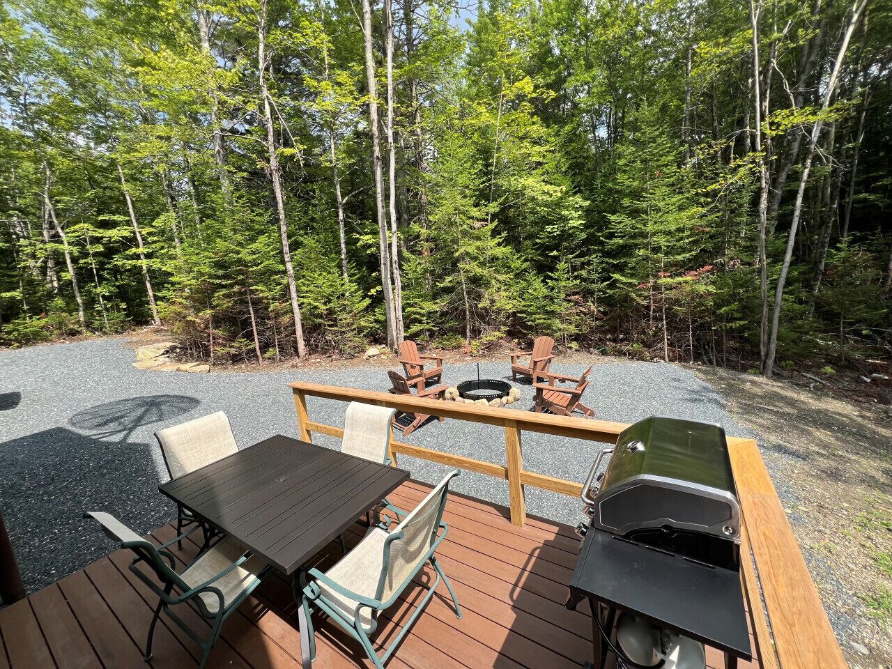 An outdoor deck with a table and chairs, a grill, and a fire pit surrounded by trees.