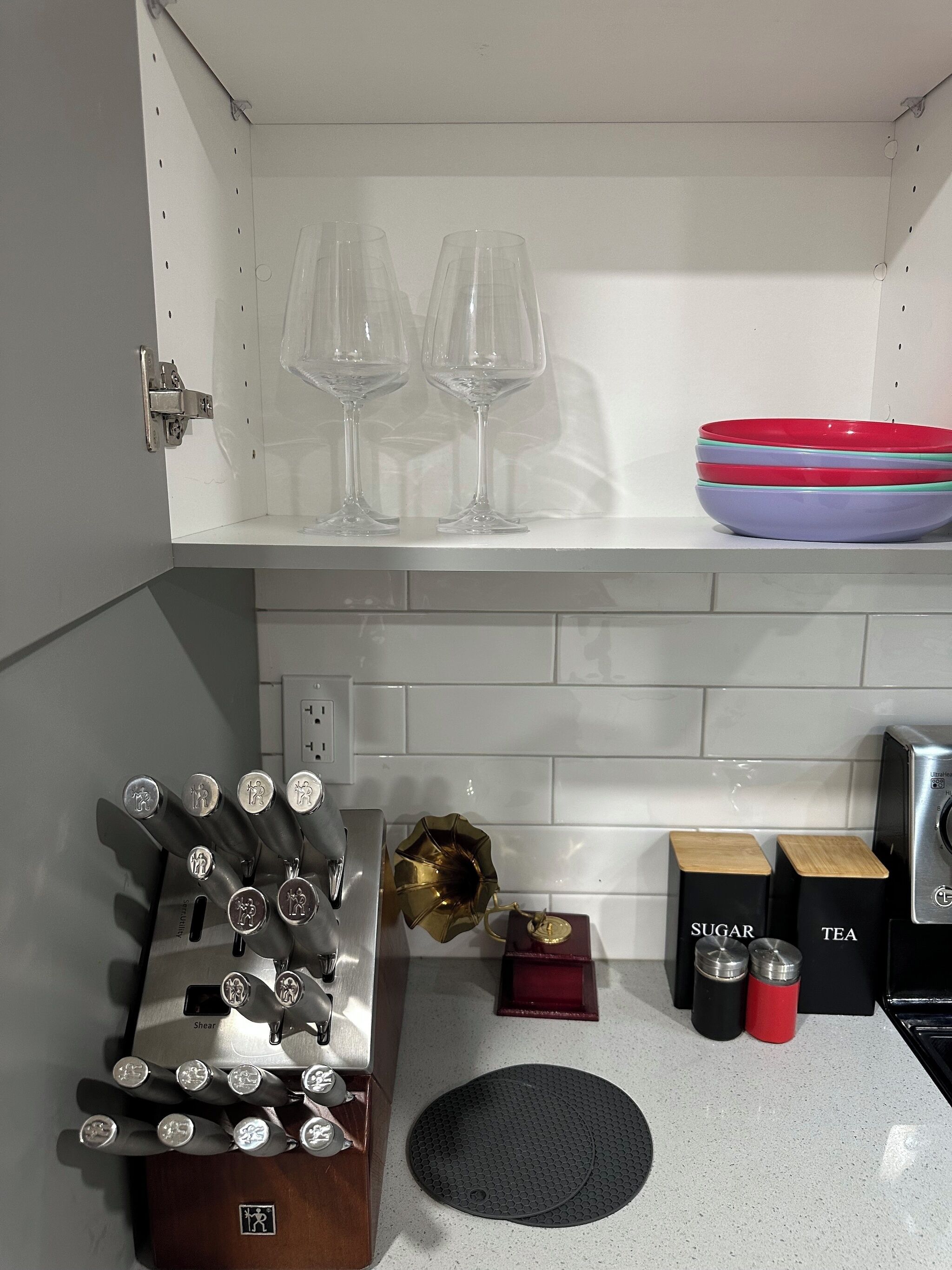 Une cuisine équipée d’un bloc à couteaux, d’une machine à café, d’un distributeur de thé et de sucre, d’un bol et de deux verres à vin placés sur une étagère.