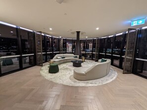 Relax in a stylish lounge with a sofa, chaise lounge, and ottomans, surrounded by floor-to-ceiling windows offering a city view.