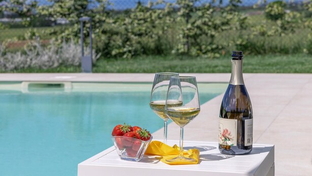 A poolside setting with a bottle of white wine, two glasses, and a bowl of strawberries.