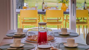 A breakfast table set with plates, cups, and a pitcher of red juice, overlooking a garden with yellow chairs.