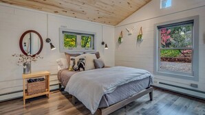 A bedroom with a wooden ceiling, a bed with a striped blanket, a bedside table with a vase of flowers, and a window with a view of trees.