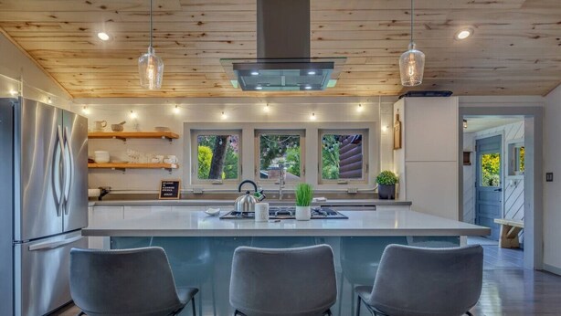 A modern kitchen with a central island, bar stools, pendant lights, and a view of the outdoors.