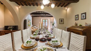 Ruokailutila, jossa on pitkä ruokapöytä katettuna, valkoiset, kuviolliset tuolit, kultareunaiset lautaset ja viinilasit.