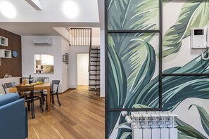 A modern living room with a wooden floor, a dining area with a wooden table and chairs, a kitchen with white cabinets, and a staircase leading to an upper floor.