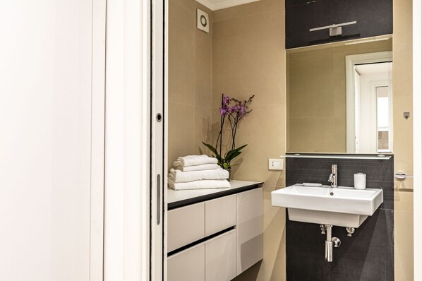 A modern bathroom with a white vanity, a sink, and a mirror. There is a stack of white towels on the vanity and a potted orchid on the counter.