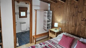 Un dormitorio con techo de madera, una cama con ropa de cama estampada, una mesita de noche con lámpara, una mesita auxiliar de madera y un armario de madera.