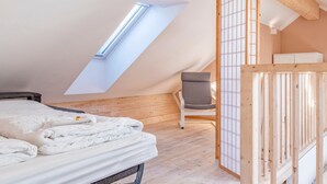Un dormitorio ático acogedor con techo inclinado, tragaluz, cama con ropa blanca, una silla gris y un armario de madera.